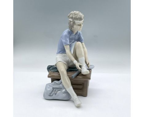 Glossy porcelain figure of a seated young tennis player with his left leg raised about to tie his shoe. Nao backstamp. Dimens