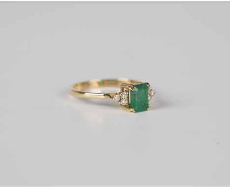 An 18ct gold, emerald and diamond ring, claw set with the cut cornered rectangular step cut emerald between circular cut diam