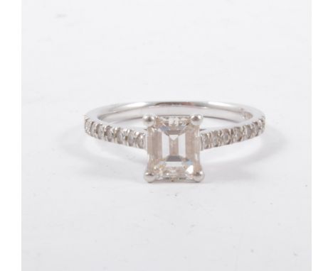 A diamond solitaire ring, the rectangular step cut stone claw set in a platinum mount with diamond set shoulders, the centre 