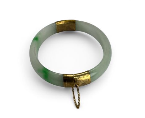 A jadeite and gilt hinged bangle. The yellow metal appears to be gilded base metal. With a safety chain, testing as circa 9ct