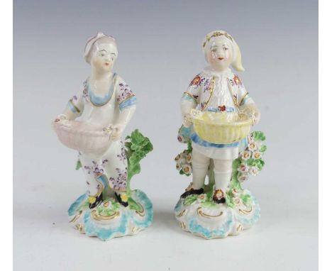 A pair of Derby porcelain figural table salts, circa 1765, each shown standing holding a basket upon a rococo base, patch mar