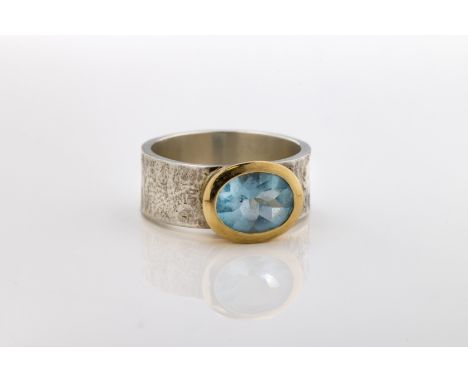 A contemporary white metal and blue topaz single stone ring, the oval mixed cut topaz in yellow gold rub over setting, hammer