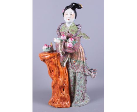 A Chinese porcelain famille rose figure of Kuan Yin holding peaches standing against a tree trunk, 15" high