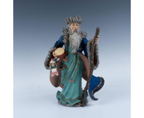 An older gentleman with a flowing beard, dressed in a teal robe and blue jacket, holds a walking stick in one hand and a drum