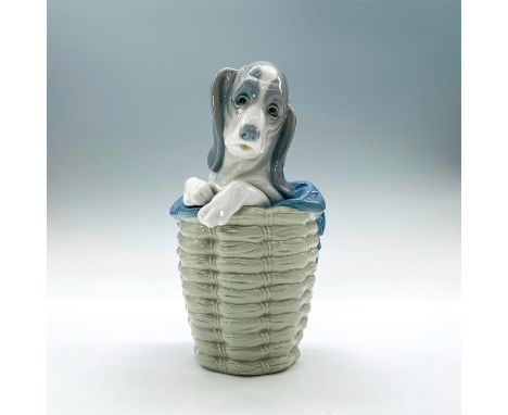 A glossy figure of a basset hound poking out of a woven laundry basket with a blue blanket wrapped around them. Lladro backst