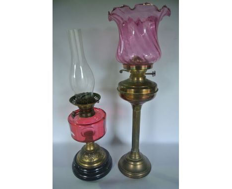A tall antique brass oil lamp on a fluted column support complete with a cranberry glass shade with shaped outline and wrythe