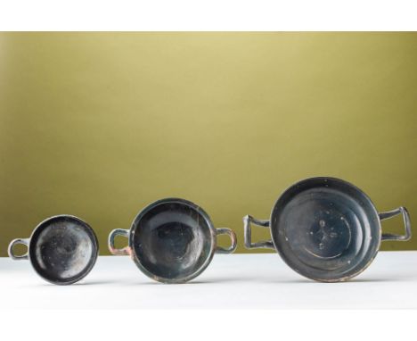 Ca. 350 - 280 BC. A group of three black-glazed ceramic cups, each with a low foot, a shallow bowl, and horizontal handles. T