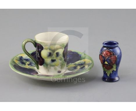 A Moorcroft 'pansy' pattern coffee cup and saucer, c.1915 and a similar later miniature vase, the coffee cup and saucer with 