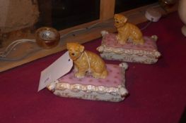 A pair of porcelain lidded boxes in the form of dogs on cushions