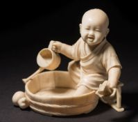 A Japanese carved ivory okimono of a young boy paddling in a wash tub: holding a toy boat in one