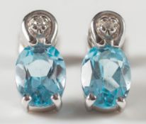 A pair of single stone, oval aquamarine ear studs:, each set with a small, circular diamond.