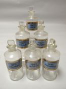 A set of six clear glass pharmaceutical bottles with stoppers, all with blue chemical labels with