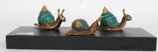Three cast metal snails having painted shells mounted on a black marble effect base