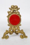 A decorative 20th century brass and velvet easel pocket watch stand adorned with cherubs on rococo