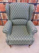 An upholstered green patterned wing back armchair raised on turned wooden legs and brass castors