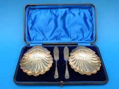 A cased pair of Victorian hallmarked silver shell shaped butter dishes and knives, Sheffield 1897,
