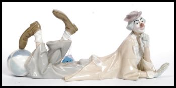 A large Lladro figure of a clown in repose leaning with his foot resting on a large circus ball.