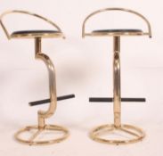 A pair of retro style gilt bar stools, having black circular seat pads and raised foot rests.