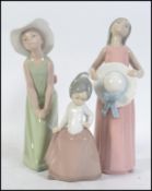 Two Lladro girls one hatted and leaning forward wearing a green dress.