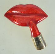 A 1980s goldtone metal and enamel brooch in the form of lips and lipstick having rollover catch.