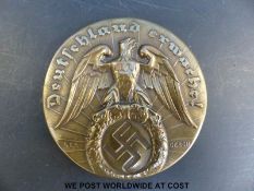 A German Nazi style brass badge with embossed emblem of an eagle carrying a swastika and script