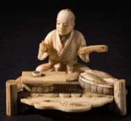 A Japanese carved ivory okimono of a scribe: seated by the riverside with a basket of fish by his