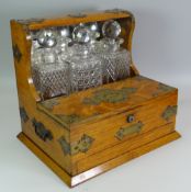 AN OAK THREE BOTTLE TANTALUS with front twin lid compartment and single drawer, brass mounted and