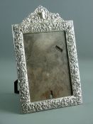 AN ELECTROPLATE EASEL PORTRAIT MIRROR, the wide frame decorated overall with flowers and scrolls