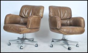 A pair of tan leather retro style swivel arm chairs raised on chrome supports with fitted castors.