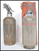 Two vintage oversized Soda Syhons by Sparklets, both with wire mesh covers and one retaining its