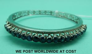 A white metal bangle set with a row of round cut sapphires with pierced design verso (21.