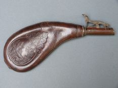 Leather and brass shot flask of curved form with embossed decoration of a man with a gun and his