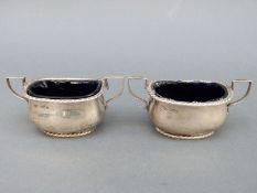 A pair of hallmarked silver two-handled open salts with blue glass liners, Sheffield 1907 and 1910,