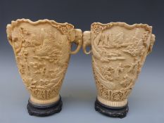A pair of faux ivory vases with carved decoration and elephant handles, on carved wooden bases,