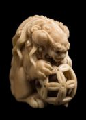 A Japanese carved ivory netsuke of a temple guardian lion-dog: holding a pierced ball containing a