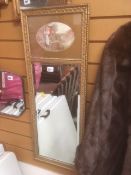 A narrow gilt framed bevelled wall mirror with print of a rustic scene to the top