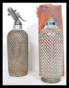 Two vintage oversized Soda Syphons by Sparklets, both with wire mesh covers and one retaining its