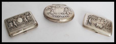 A collection of 3 white metal cigarette cases all pressed in relief with village scenes. One dated