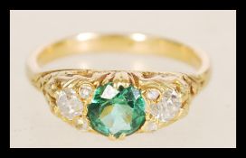 A gold ring with a central green tourmaline stone in decorative floral settings flanked by two 20
