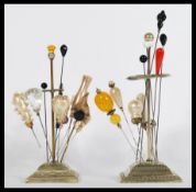 Two vintage white metal square based hat pin stands containing a collection of vintage hat pins