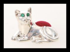 A silver pincushion in the form of a cat having green stone eyes and a red cushion to its back.