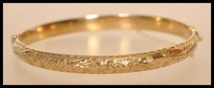 A hallmarked 9ct gold bangle with a hinged opening and safety chain with floral engraving.