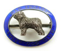 A silver brooch set with an Old English Sheep Dog surrounded by blue enamel reading 