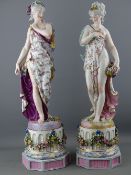 A PAIR OF CONTINENTAL CHINA STANDING FLORAL BEDECKED FEMALE FIGURINES, 46 cms high, standing atop