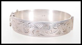 A hallmarked silver bangle with scrolled engraving and a hinge opening with safety chain. Hallmarked