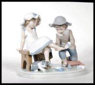 A Lladro ceramic figure group titled ' Try this One ', (5361) of a young girl trying on a shoe.