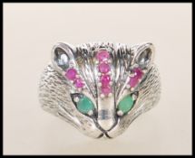 A silver ring in the form of a cats head with emerald eyes and channel set ruby's to the head.