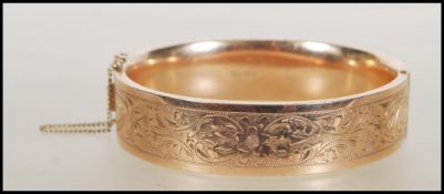 A 9ct gold bangle. The bangle having foliate decoration and hidden clip clasp with safety chain.