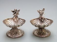 A pair of novelty white metal salts formed as shell shaped bowls with cherubs playing instruments,