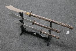 Three graduated Chinese style twin handled swords on a wooden stand